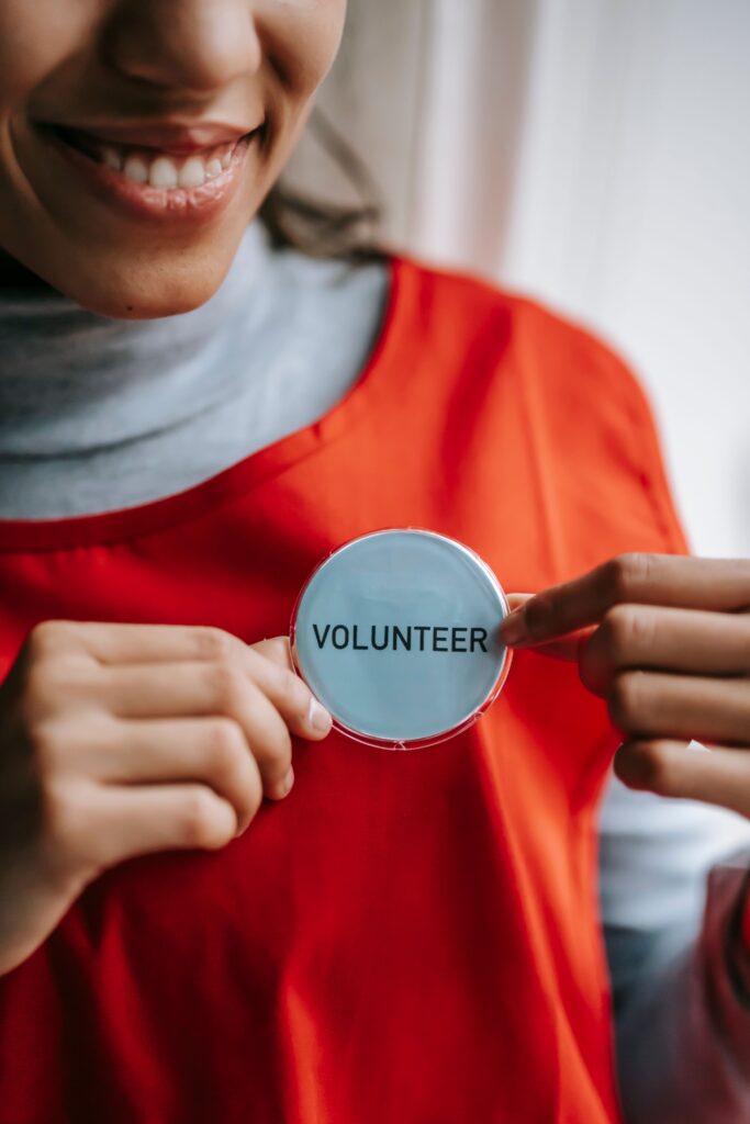pexels photo 6348129 6348129 Crop anonymous positive ethnic female smiling and demonstrating sign with inscription volunteer on bright red apron